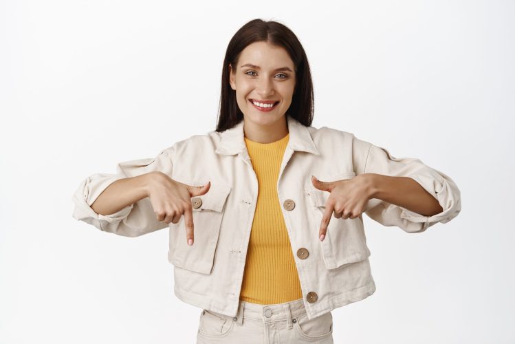 Image of smiling candid woman, pleasant friendly face expression, pointing fingers down, showing sale advertisement below, standing over white background. Copy space