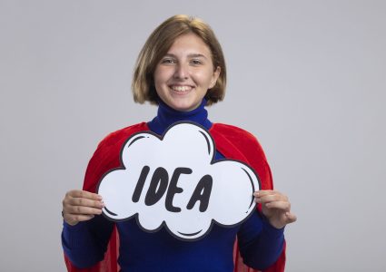 smiling young blonde superhero girl in red cape holding idea bubble looking at camera isolated on white background with copy space